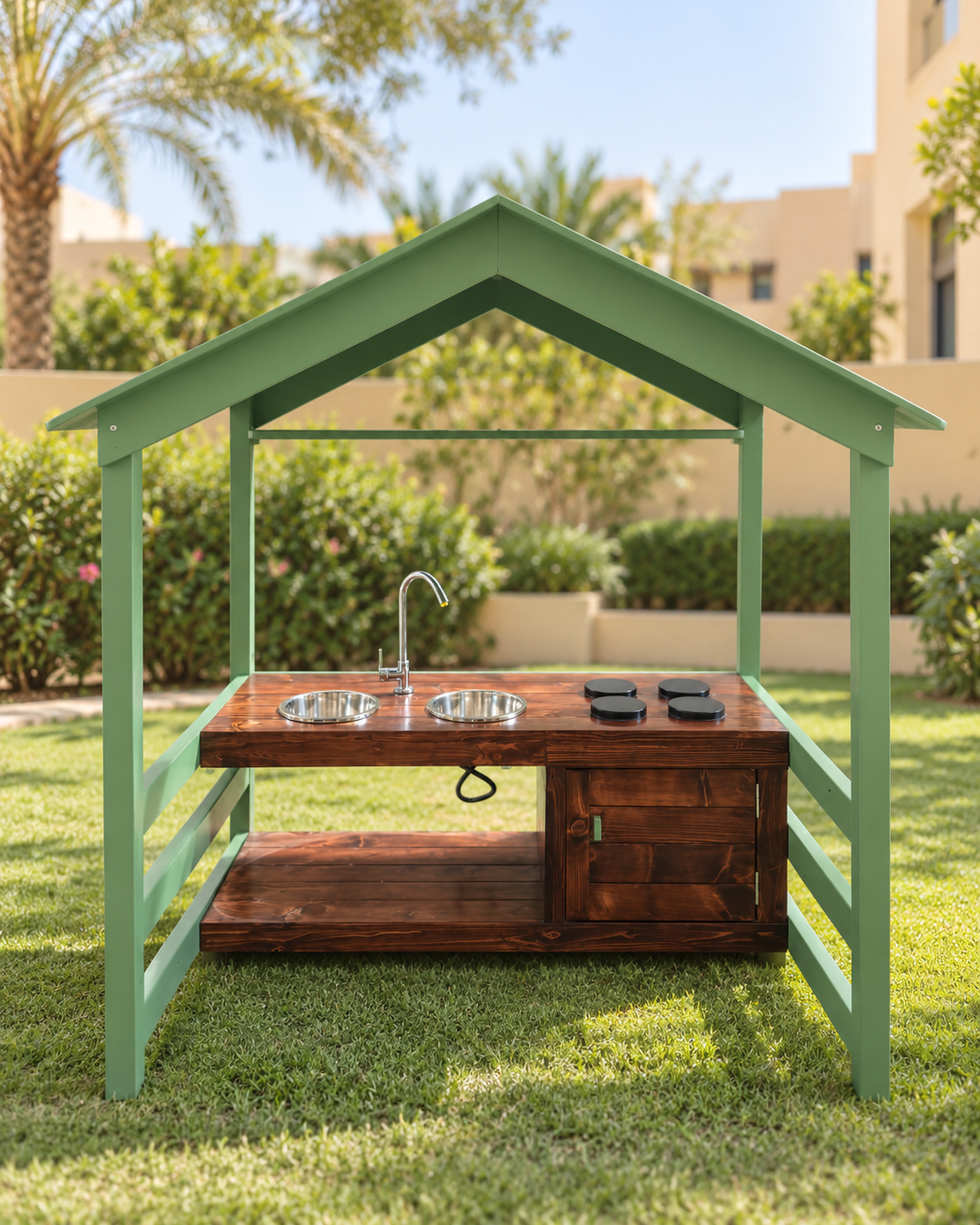 Outdoor play kitchen set with a green house-shaped frame and a dark brown wooden countertop featuring two stainless steel sinks, a faucet, and cookware, situated on a green lawn.