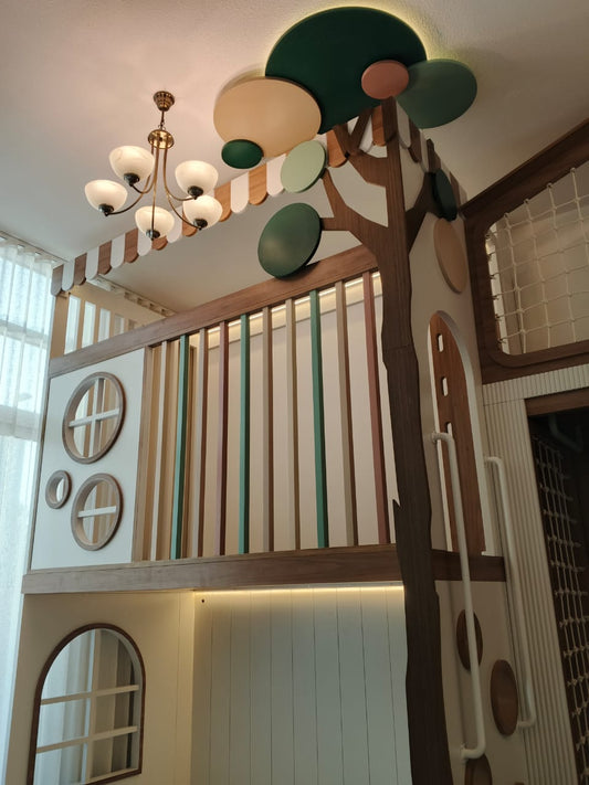 A whimsical children's loft play area featuring a tree-themed climbing structure with a brown trunk, green and beige circular tree tops, and a striped canopy. The loft has a white paneled upper level with a slatted wooden railing, circular window cutouts, and warm under-cabinet lighting.