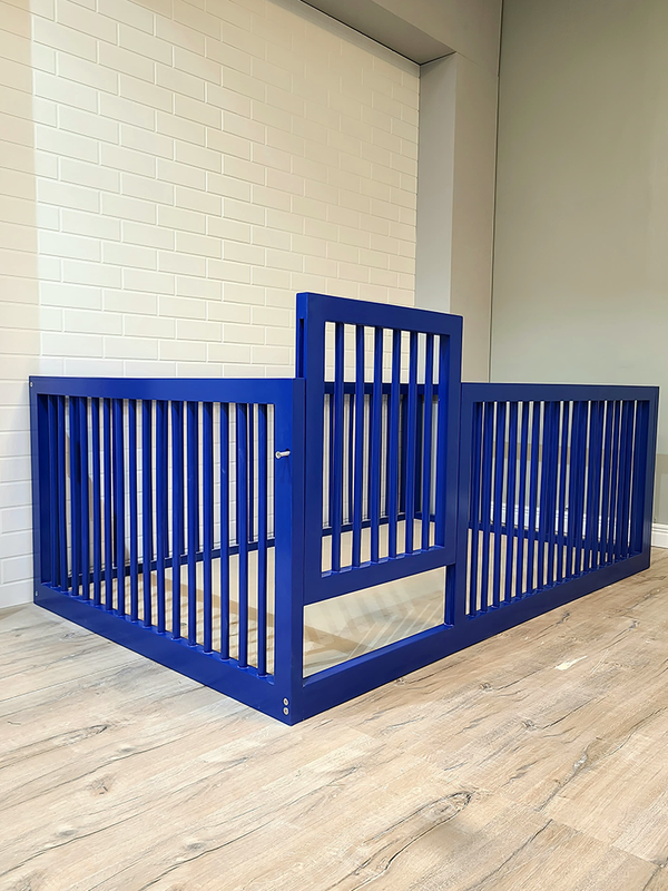 A blue metal bed frame with vertical rails, positioned on a wooden floor against a white brick wall background.