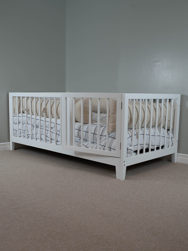 A white toddler bed with a striped bedsheet, positioned on a carpeted floor against a plain wall.