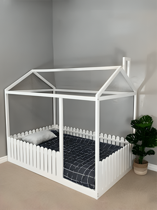 A white bed frame designed to look like a picket fence with a transparent roof, positioned in a room with a potted plant beside it.