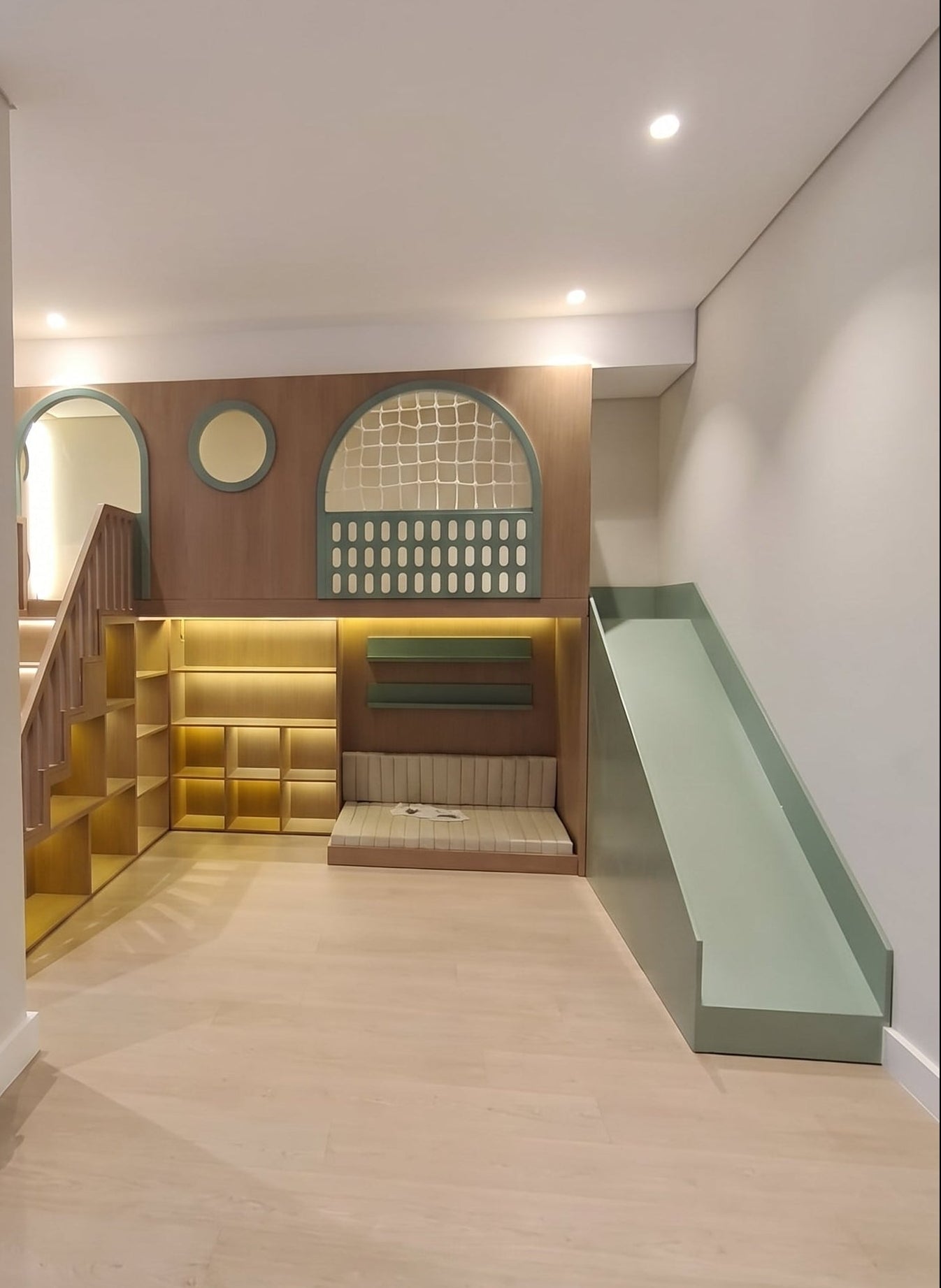 A modern children's playstructure featuring a green slide, underneath seating, shelves bith built-in LED light.