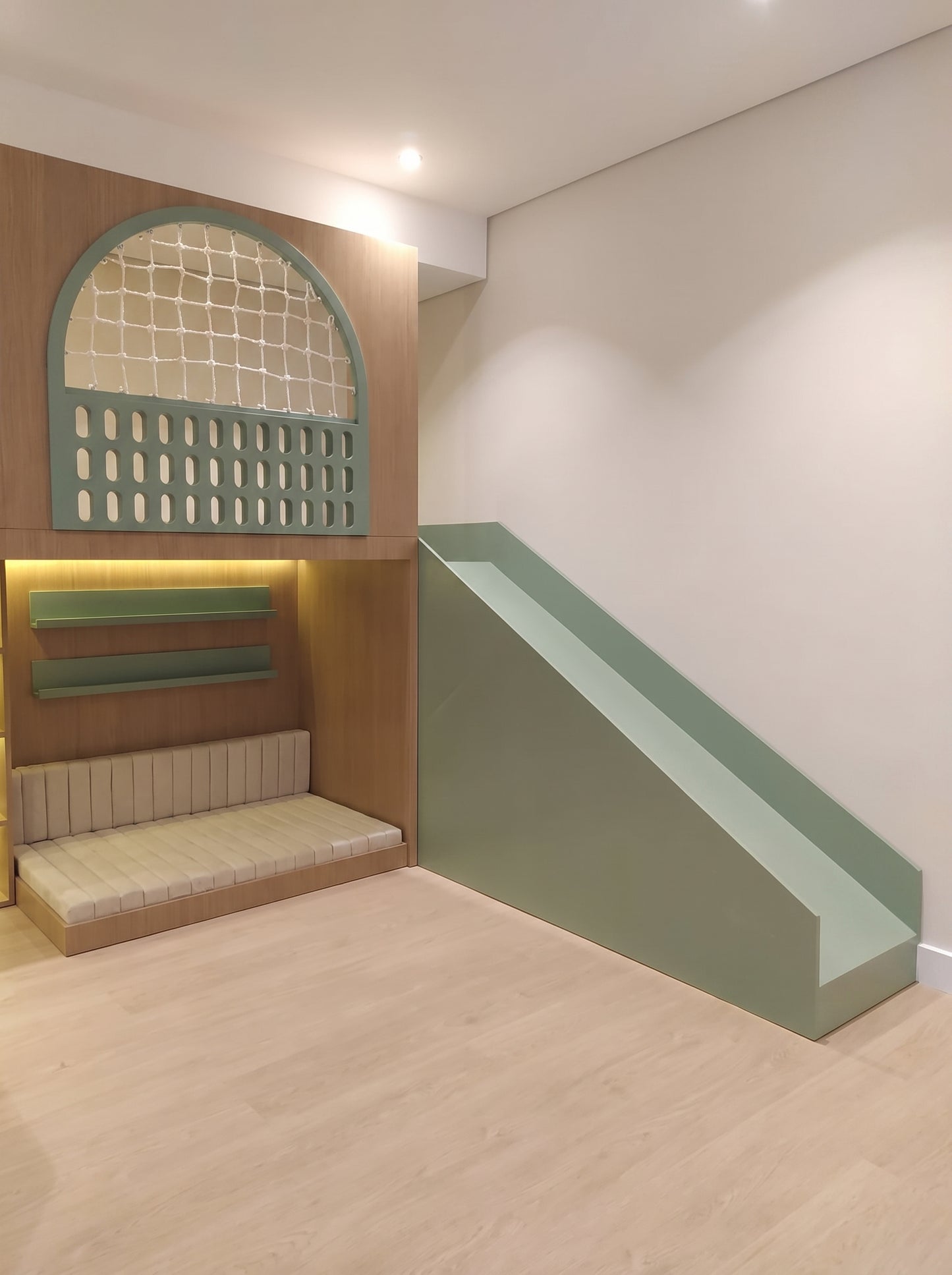 A modern children's playroom corner featuring a built-in wooden bed with a green arched window, shelves and seating, and a green slide.