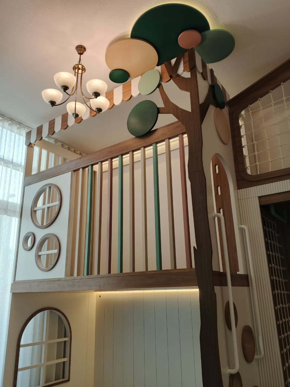 A whimsical children's loft play area featuring a tree-themed climbing structure with a brown trunk, green and beige circular tree tops, and a striped canopy. The loft has a white paneled upper level with a slatted wooden railing, circular window cutouts, and warm under-cabinet lighting.