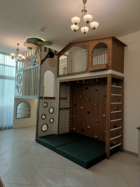 A large wooden children's play structure in a playroom, featuring a loft bed with a climbing wall, ladder, and green padded floor mats. The room has tiled floors, white walls, and two hanging pendant lights.