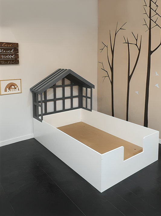 A twin-sized bed with a white frame designed to resemble a house with a gray roof and window frames, positioned in a room with a beige wall and tree decals.