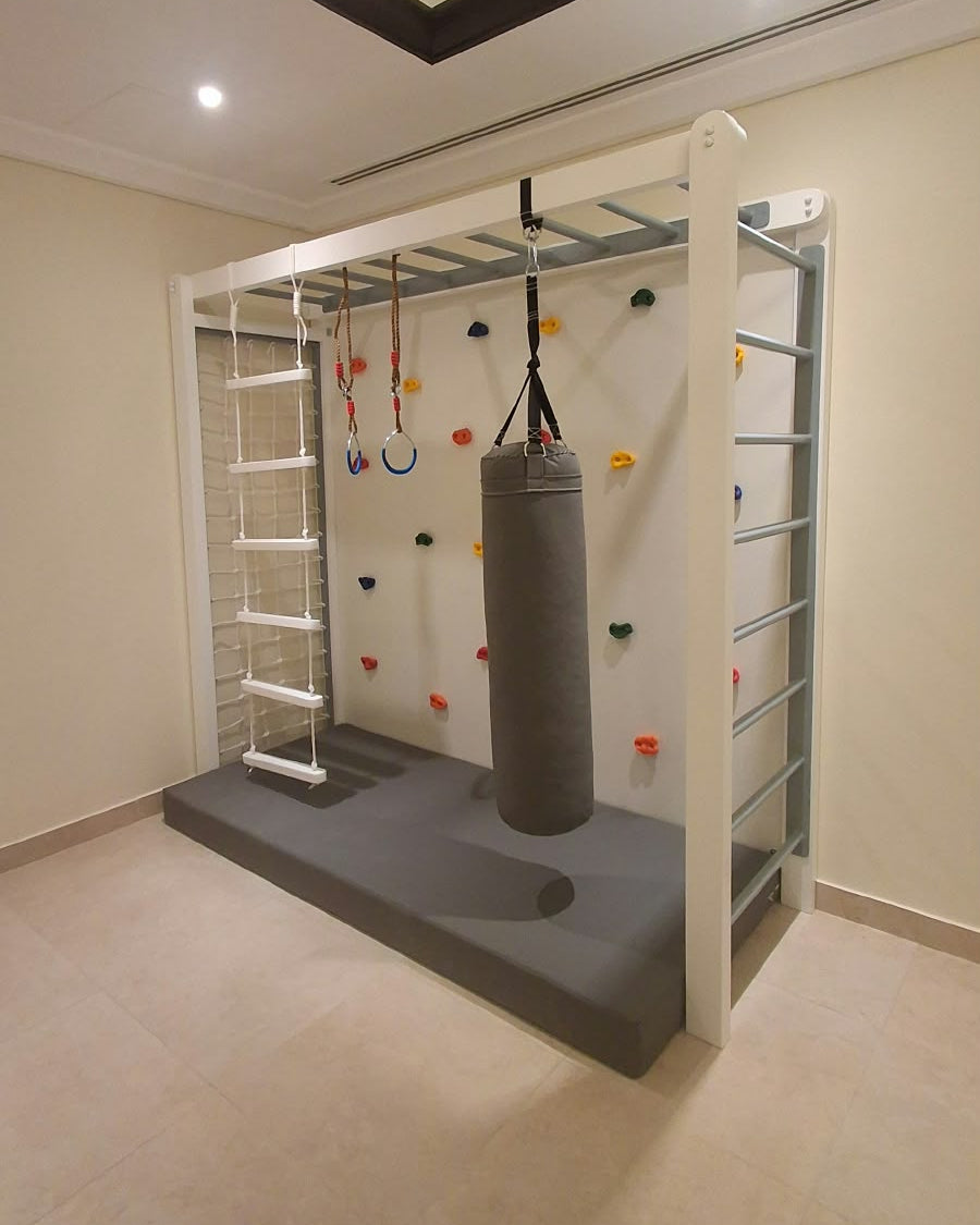 kids indoor gym with climbing wall having colorful climb holds, monkey bars, rope ladder, punching bag, gym rings, spider rope net, and safety mat.