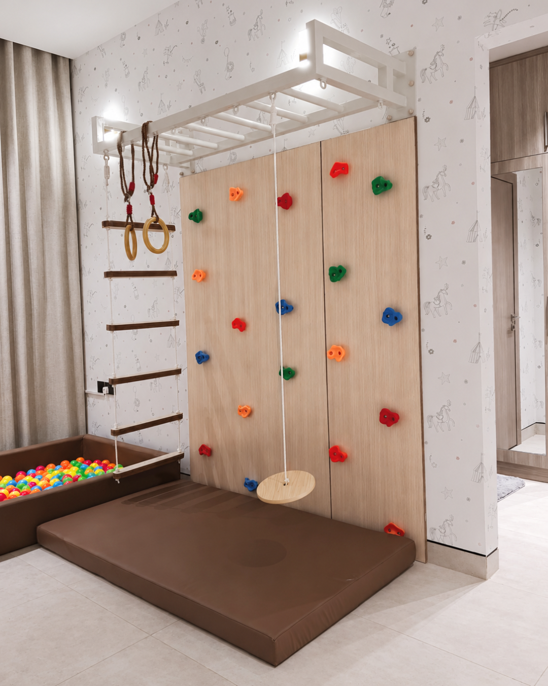 A children's indoor climbing wall with colorful handholds, a white monkey bar, hanging gym rings, a disc swing, a rope ladder, a brown padded floor mat, and a ball pit in a bright room.