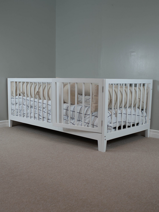 A white toddler bed with a striped bedsheet, positioned on a carpeted floor against a plain wall.
